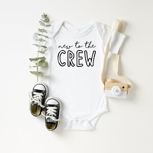 May include: White baby onesie with the text "new to the CREW" in black. The onesie is paired with small black and white sneakers, a wooden toy camera, and greenery. A cute baby outfit.
