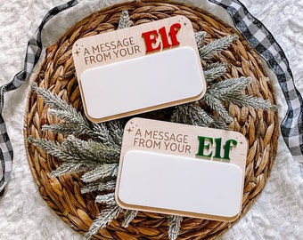May include: Two wooden message boards with the text "A MESSAGE FROM YOUR Elf" in red and green. The boards have a white rectangular space for writing and are on a woven basket with greenery and a black and white ribbon.