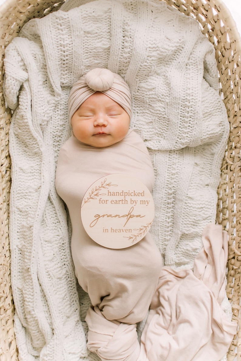 Peut inclure: Un nouveau-n&eacute; envelopp&eacute; dans un lange beige, portant un turban beige, est couch&eacute; dans un panier en osier. Une pancarte en bois avec le texte "handpicked for earth by my grandpa in heaven" est plac&eacute;e sur la poitrine du b&eacute;b&eacute;.