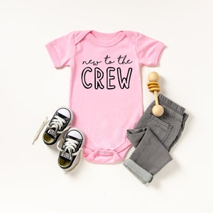 May include: A pink baby onesie with the text "new to the CREW" in black. The onesie is paired with gray pants, black and white sneakers, and a wooden rattle. The outfit is laid out on a white surface.