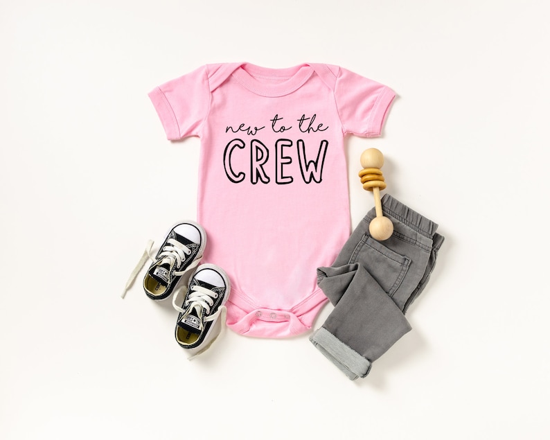 May include: A pink baby onesie with the text "new to the CREW" in black. The onesie is paired with gray pants, black and white sneakers, and a wooden rattle. The outfit is laid out on a white surface.
