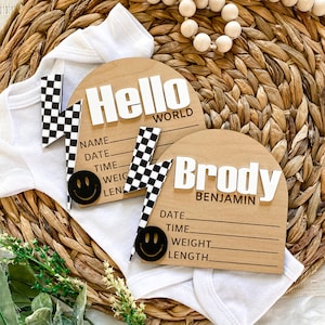 May include: Two wooden baby announcement plaques with a checkered racing flag design. The top plaque says "Hello World" and the bottom plaque says "Brody Benjamin". Both plaques have spaces to fill in the baby's name, date, time, weight, and length.