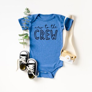 May include: A blue baby onesie with the text "new to the CREW" in black. The onesie is surrounded by a sprig of greenery, a pair of black and white baby sneakers, and a small wooden toy camera.