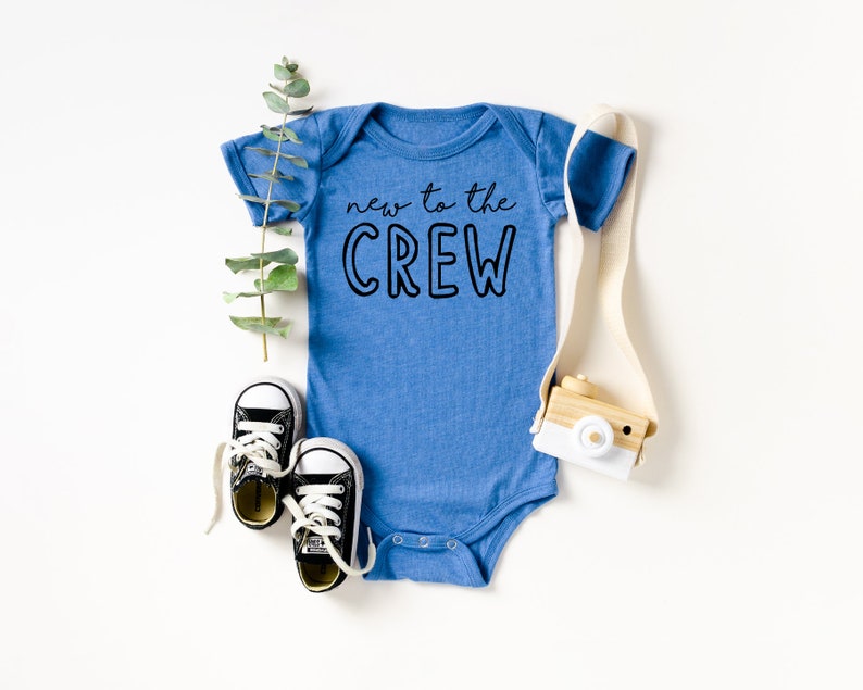 May include: A blue baby onesie with the text "new to the CREW" in black. The onesie is surrounded by a sprig of greenery, a pair of black and white baby sneakers, and a small wooden toy camera.