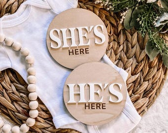 Könnte beinhalten: Zwei runde Holzschilder mit den Worten "She's Here" und "He's Here" in weißen Buchstaben auf einem hellbraunen Hintergrund.