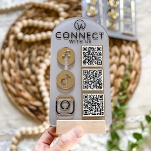 Peut inclure: Un panneau en acrylique gris en forme de demi-cercle avec le texte CONNECT WITH US et un logo en haut. Il présente trois icônes de réseaux sociaux dorées à gauche et trois codes QR carrés correspondants à droite, fixés sur un socle en bois clair.