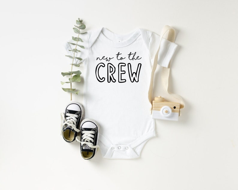 May include: White baby onesie with the text "new to the CREW" in black. The onesie is paired with small black and white sneakers, a wooden toy camera, and greenery. A cute baby outfit.