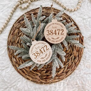 May include: Two wooden Christmas ornaments with the text "THERE'S NO PLACE LIKE home 38472 FOR THE HOLIDAYS" are displayed. The ornaments are round with a string for hanging. They are surrounded by frosted evergreen branches and rest on a woven mat.