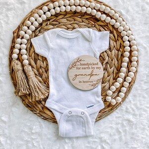 Peut inclure: Un body blanc pour b&eacute;b&eacute; avec une pancarte en bois qui dit "handpicked for earth by my grandpa in heaven". Le body est sur un panier en osier avec un brin de perles en bois.