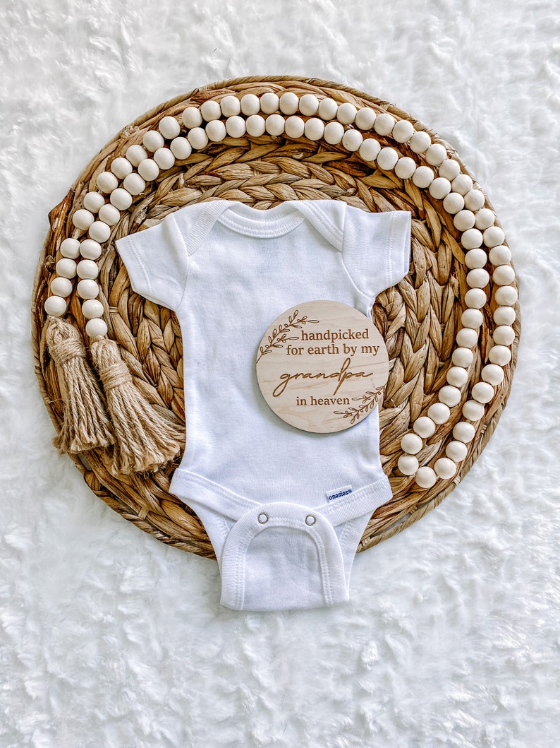 Peut inclure: Un body blanc pour b&eacute;b&eacute; avec une pancarte en bois qui dit "handpicked for earth by my grandpa in heaven". Le body est sur un panier en osier avec un brin de perles en bois.