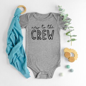 May include: A gray baby onesie with the text "new to the CREW" in black. A blue swaddle blanket, wooden toy, and eucalyptus sprig are arranged nearby. Small, colorful felt balls add a playful touch to the scene.