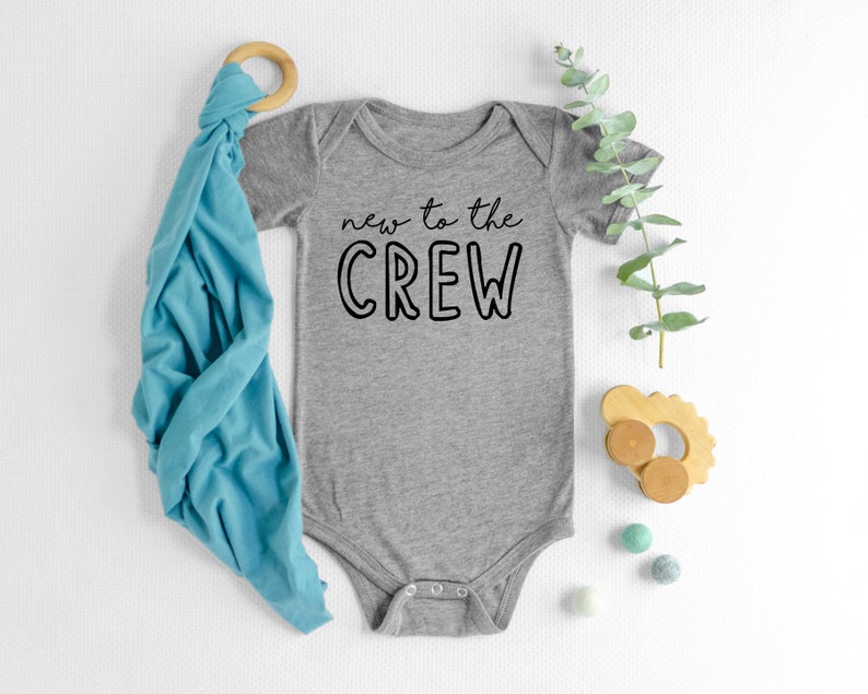 May include: A gray baby onesie with the text "new to the CREW" in black. A blue swaddle blanket, wooden toy, and eucalyptus sprig are arranged nearby. Small, colorful felt balls add a playful touch to the scene.