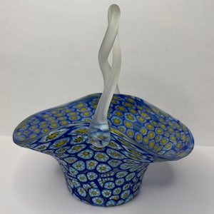 May include: A glass basket featuring a millefiori pattern with blue, white, and yellow floral motifs. The bowl has a wide, ruffled rim and a single, tall, twisted handle made of clear, frosted glass that arches over the center of the basket.
