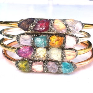 May include: A set of four gold, silver, and copper bracelets with different colored gemstones. Each bracelet features a row of small, rough-cut gemstones set in a metal band.