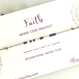 Puede incluir: Una tarjeta blanca con la palabra "Faith" en escritura rosa y el texto "Morse Code Bracelet" en escritura rosa más pequeña. La tarjeta también incluye el texto "International Morse Code" y el código Morse de la palabra "Faith". Un brazalete de cuerda marrón con cuentas blancas y azules se muestra en la tarjeta.