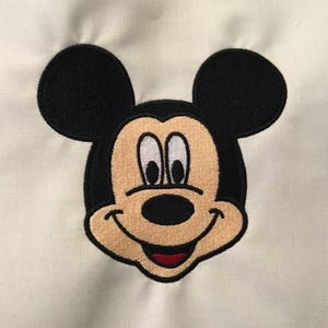 May include: Embroidered patch of Mickey Mouse's face with black ears and a white background.