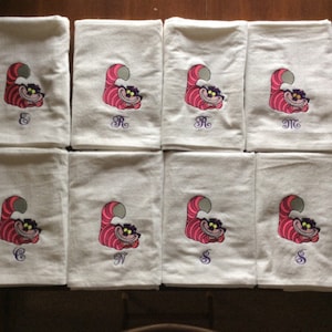 May include: Eight white kitchen towels with embroidered images of the Cheshire Cat from Alice in Wonderland. Each towel has a different initial embroidered in purple.