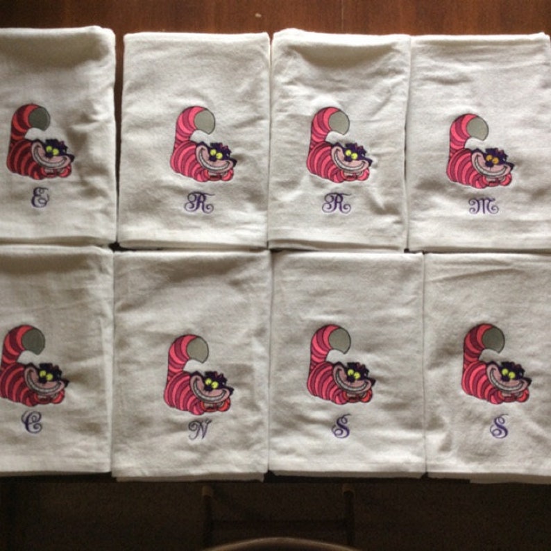 May include: Eight white kitchen towels with embroidered images of the Cheshire Cat from Alice in Wonderland. Each towel has a different initial embroidered in purple.