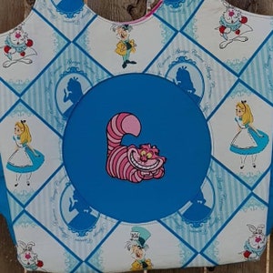 May include: A white fabric tote bag with a blue circle in the centre. The circle features a pink and white Cheshire Cat from Alice in Wonderland. The bag is decorated with a checkered pattern and various Alice in Wonderland characters, including Alice, the Mad Hatter, and the White Rabbit. The text "Curiouser and Curiouser" is repeated around the circle.
