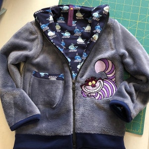 May include: A blue fleece zip-up jacket with a pocket on the left side. The jacket has a blue and white whale print lining and a purple and white Cheshire Cat appliqu&eacute; on the front.