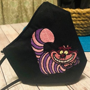 May include: Black fabric face mask featuring an embroidered Cheshire Cat from Alice in Wonderland, with purple and pink stripes.