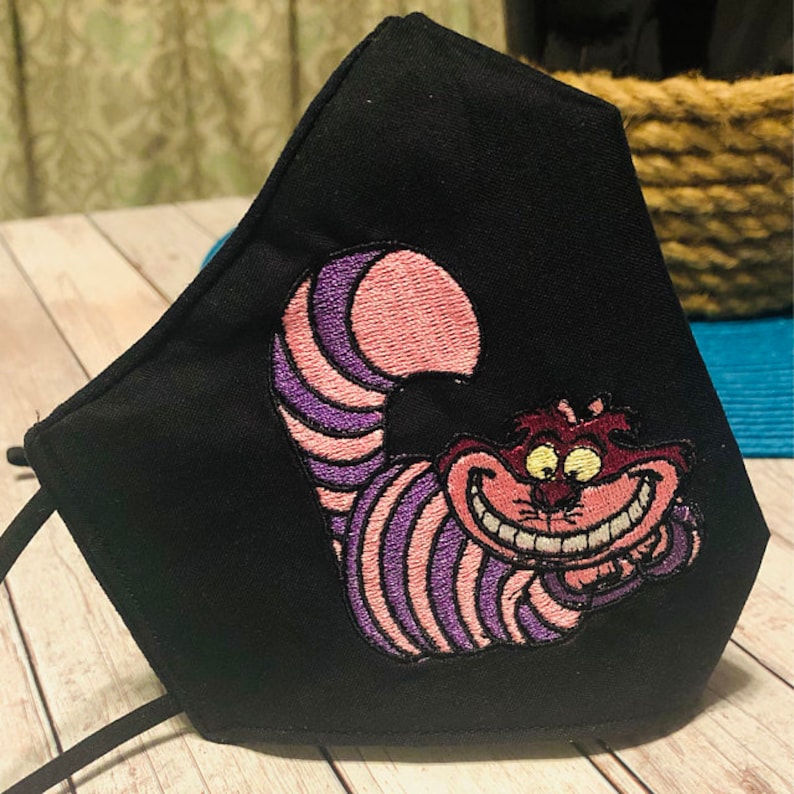 May include: Black fabric face mask featuring an embroidered Cheshire Cat from Alice in Wonderland, with purple and pink stripes.