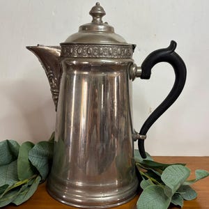 May include: A vintage silver-colored coffee pot with a black handle. The pot has a decorative rim and a lid with a small finial. The pot is sitting on a wooden surface with green leaves.
