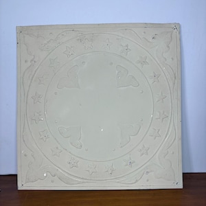 May include: Off-white square decorative metal tile with embossed star and heart designs. A central circle features four heart shapes and is surrounded by stars. The corners have a floral pattern. The tile is likely for interior design.