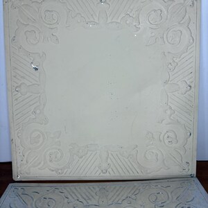 May include: Two square, off-white decorative tiles with ornate, raised floral and scrollwork borders. The top tile has a cream-colored surface, while the bottom tile is a metallic silver color. The tiles are likely for use in home decor or craft projects.