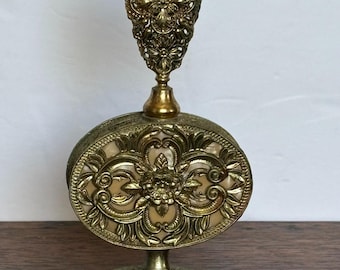May include: A gold metal perfume bottle with a decorative, ornate design. The bottle has a round body with a filigree pattern and a tall, pointed stopper. The bottle sits on a matching stand with a filigree base.