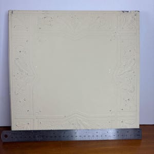 May include: Off-white square ceiling tile with embossed floral and geometric patterns. A metal ruler is placed along the bottom edge. The tile has a decorative border with ornate designs and a central plain area.