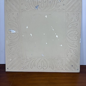 May include: A square, off-white tile with an embossed floral and scroll design. The tile has a central, plain square surrounded by a border with corner squares and diagonal lines. The design is symmetrical and features a vintage aesthetic.