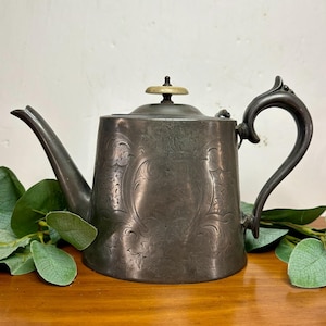May include: Antique pewter teapot with a decorative floral pattern. The teapot features a curved spout, a curved handle, and a lid with a small knob. The teapot is a dark gray color and sits on a wooden surface.