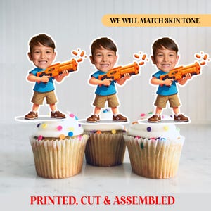 May include: Three cupcakes topped with white frosting and colorful sprinkles. Each cupcake has a cutout of a boy holding an orange Nerf gun. Text at the top says "WE WILL MATCH SKIN TONE" and at the bottom "PRINTED, CUT & ASSEMBLED."