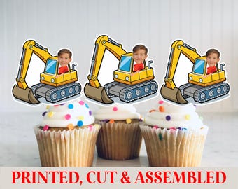 May include: Three cupcakes topped with white frosting and colorful sprinkles, each adorned with a yellow excavator cutout featuring a child's face. The text "PRINTED, CUT & ASSEMBLED" is at the bottom.
