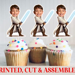 May include: Three cupcake toppers featuring a cartoon boy with a lightsaber, above three cupcakes with white frosting and colorful sprinkles. The text "PRINTED, CUT & ASSEMBLED" is at the bottom.