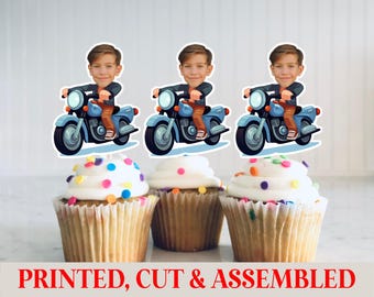 May include: Three cupcakes topped with white frosting and colorful sprinkles. Each cupcake has a decorative topper featuring a cartoon boy riding a blue motorcycle. The text "PRINTED, CUT & ASSEMBLED" is at the bottom.