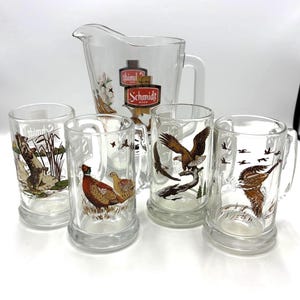 May include: A clear glass pitcher and four matching beer mugs. The pitcher and mugs feature wildlife illustrations, including birds and a hunting scene. The pitcher has the "Schmidt Beer" logo. The mugs have handles.