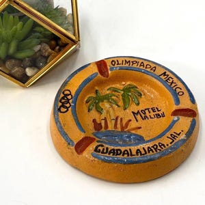 May include: A round, yellow ceramic ashtray featuring a hand-painted palm tree scene, the text "OLIMPIADA MEXICO", "MOTEL MALIBU", and "GUADALAJARA, JAL.", along with the Olympic rings symbol. The rim has blue accents and three dark red notches.