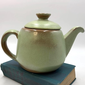 May include: A ceramic teapot with a rounded body, a curved handle, and a spout. The teapot features a matte light green glaze with brown accents along the rim, handle, spout, and lid. The lid has a small knob handle at the top.