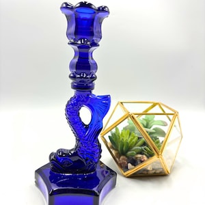 May include: A cobalt blue glass candlestick with a mermaid design. The candlestick has a fluted top and a round base.