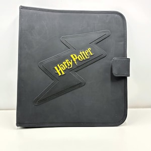 May include: Black Harry Potter binder with a yellow lightning bolt and text. The binder has a snap closure and is made of a textured material. The Harry Potter logo is in yellow.