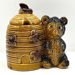 May include: A ceramic honey pot shaped like a beehive with a bear figurine. The honey pot is brown with a lid and bee accents. The bear is brown and black with a red nose. The honey pot and bear are on a white surface.
