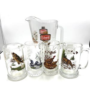 May include: A clear glass pitcher and four matching beer mugs. The pitcher and mugs feature nature-themed illustrations of a fish, ducks, turkeys, and a deer. The pitcher has a white handle and a Schmidt Beer label.