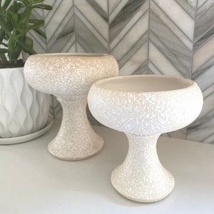 May include: Two off-white, textured pedestal bowls with a speckled finish. The bowls have a wide, shallow top and a rounded base, perfect for displaying small plants or decorative items. The bowls are placed on a white surface.
