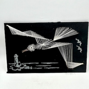 May include: Black string art depicting a seagull flying over a lighthouse and waves. The seagull is made of white string on a black background.