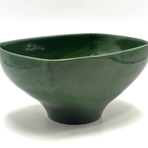May include: A dark green ceramic bowl with a square top and a rounded, flared base. The bowl has a glossy finish and is set against a white background. The bowl's shape is modern and minimalist.