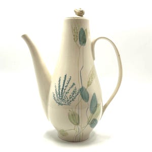 May include: Cream-colored ceramic coffee pot with a long spout and handle. The pot is decorated with a botanical design in shades of blue and green. The lid has a small, round knob.
