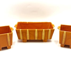 May include: Three orange ceramic rectangular planters with a striped design. Each planter has a small footed base and handles on the sides. The planters are arranged on a white surface.