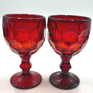 May include: Two matching ruby red glass goblets featuring a prominent embossed honeycomb or dot pattern across the bowl. Each glass has a thick, faceted stem and a wide, circular base, showcasing a deep, translucent red color throughout.